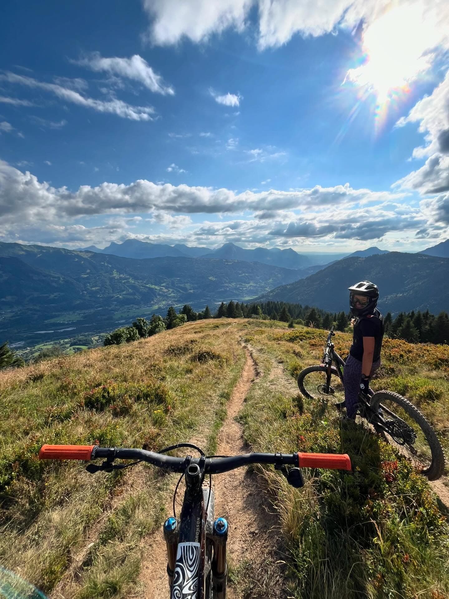 A mountain biker pausing on a ridgeline trail in the Alps, with the valley spread out below
