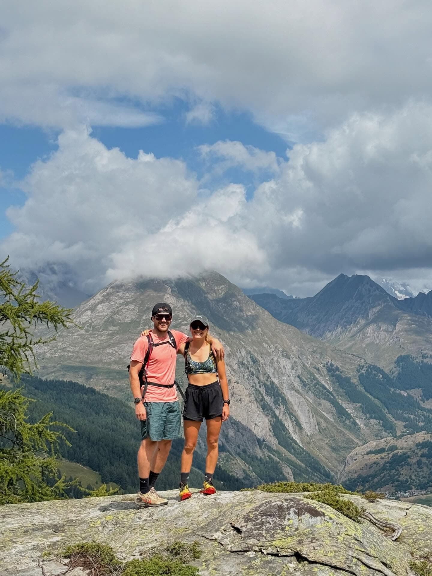 Will and Issy on a mountain summit, arms around each other, with the Alps behind them
